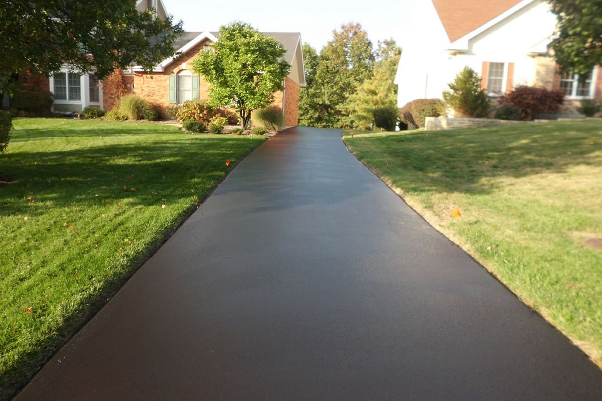 Freshly paved asphalt driveway surrounded by green lawns and trees, leading to brick houses in a suburban neighborhood under a clear sky.