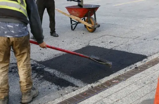 Workers repairing a small section of pavement with asphalt. One worker smooths the asphalt with a tool, while a red wheelbarrow stands nearby. The pavement shows signs of previous wear and patching.