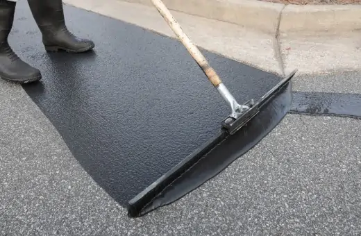 A person in black boots uses a squeegee to spread fresh asphalt or tar on a road next to a curb. The new surface appears dark and smooth in contrast to the older, lighter gray pavement.