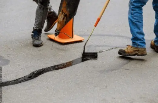 Two workers repairing a pavement crack. One is pulling tar with a shovel, standing near an orange traffic cone. The other is using a hand tool to apply sealant. Both wear jeans and boots. The ground appears to be gray asphalt.