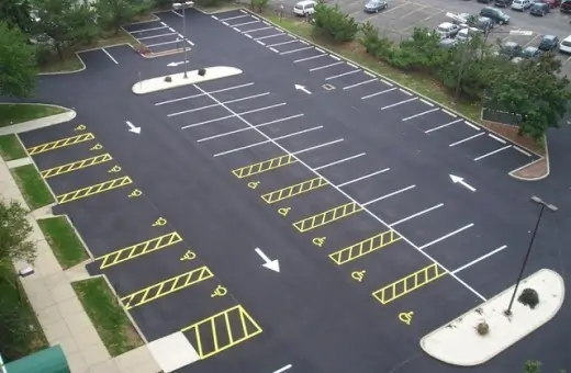 An aerial view of a freshly paved, empty parking lot with clearly marked white lines and yellow-striped handicap spaces. The lot is surrounded by greenery, and a small section of another parking area is visible in the background.