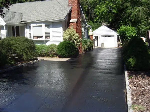 A freshly paved driveway leads to a detached garage behind a house. The house has a sloped roof, a red brick chimney, and white siding. Bushes and greenery line the driveway, and trees are visible in the background.