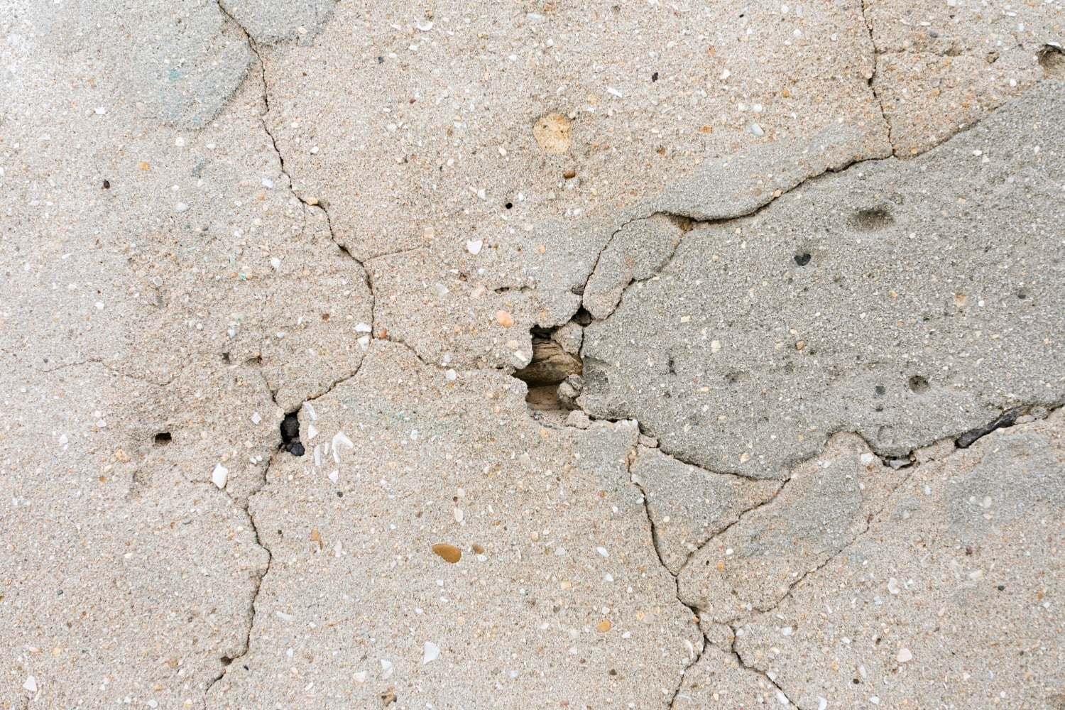 Close-up of a cracked concrete surface, showing various sizes of cracks radiating from a central point. The texture is rough, with small pebbles and uneven surfaces visible within the fissures. The color is a mix of gray and beige.