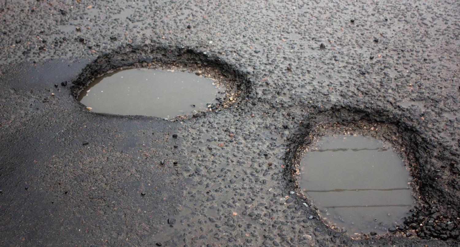 Two potholes filled with rainwater on an asphalt road. The rough, textured surface surrounds the water-filled holes, indicating recent rainfall. The uneven surface contrasts with the smooth reflection of water in the potholes.