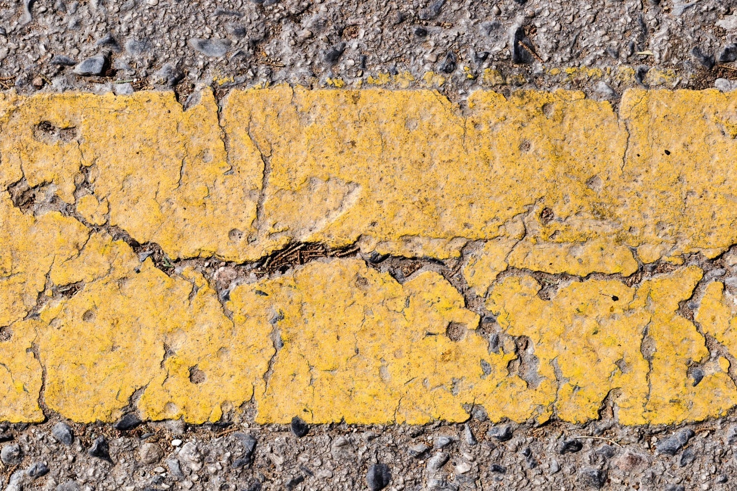 Close-up of a cracked and weathered yellow road line on asphalt. The paint is chipped and worn, revealing rough textures and small pebbles embedded in the concrete surface.
