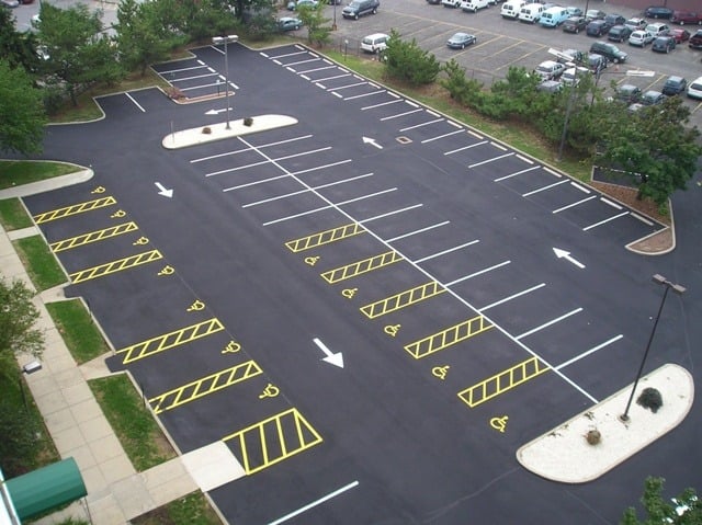 Aerial view of an empty parking lot with freshly painted white and yellow lines. Several designated handicapped spaces are visible. The lot is surrounded by trees and there are more parked cars in the distance.