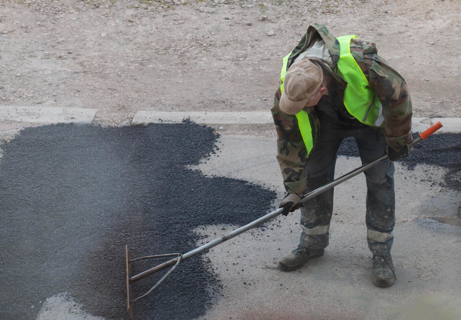 Worker in a high-visibility vest and camouflage jacket uses a rake to spread asphalt on a road. The asphalt is dark and freshly laid. The background shows a dirt surface.