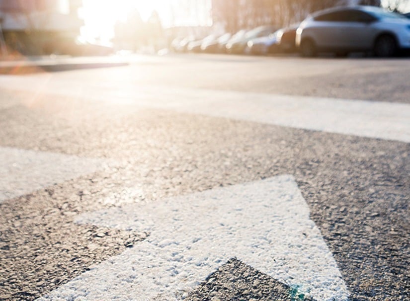 Close-up of a white arrow painted on an empty street, pointing forward. The sun is shining brightly, creating a warm glow. Several parked cars line the right side of the street in the background.