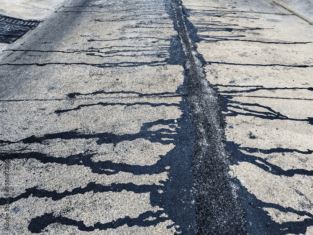 Close-up of a cracked concrete road with black tar filling the central crack. Dark streaks radiate from the repaired area across the light gray surface, creating a striking contrast. The texture of the concrete is weathered and rough.