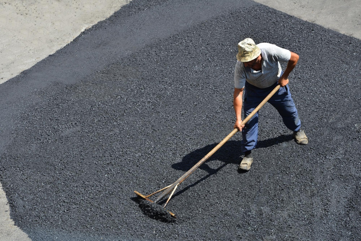 A worker in a gray shirt and blue pants spreads freshly laid asphalt with a large rake. He is wearing a beige hat and positioned on a section of smooth, black asphalt, surrounded by unpaved ground.
