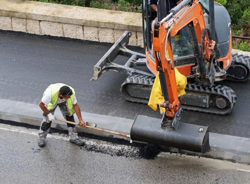 A construction worker in a vest uses a tool to smooth asphalt on a road, while a small orange excavator assists nearby. The scene takes place beside a stone wall and greenery.